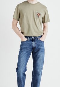 Olivgrön bomull t-shirt med en tigervarför på vänster bröst, i kombination med blå denimjeans, stående mot en neutral bakgrund.