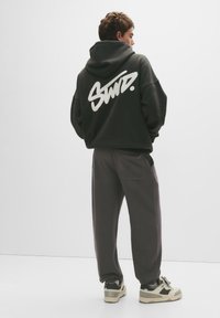 Zwarte oversized hoodie met witte grafische tekst op de achterkant, gecombineerd met donkergrijze joggers en beige sneakers met zwarte accenten.