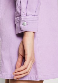 Lila Jeansjackenmanschette mit einem silbernen Knopf und gestickten Details. Eine Hand ist sichtbar, die auf dem Stoff ruht und die Textur sowie die Passform zeigt.