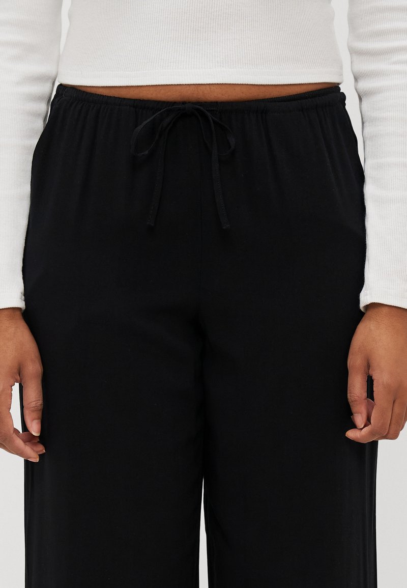 Pantalons larges noirs en tissu doux. Dotés d'une taille élastique avec cordon de serrage. Texture lisse, sans motifs ni accents visibles.