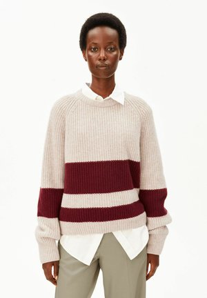 Femme aux cheveux courts et bouclés porte un pull en maille beige avec deux larges bandes horizontales bordeaux et une chemise blanche à col en dessous.