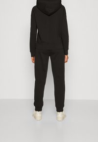 Felpa nera con cappuccio e joggers abbinati, realizzati in un tessuto morbido. Presenta una tasca a marsupio e polsini sui polsi e alle caviglie. Sneakers di colore chiaro.