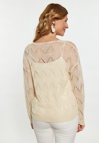 Lys beige strikket sweater med et zigzag-mønster, åben vævning og lange ærmer, parret med hvide bukser.