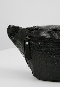 Banane en similicuir noir effet crocodile, poche zippée avant et tirette en tissu sur fond gris uni.