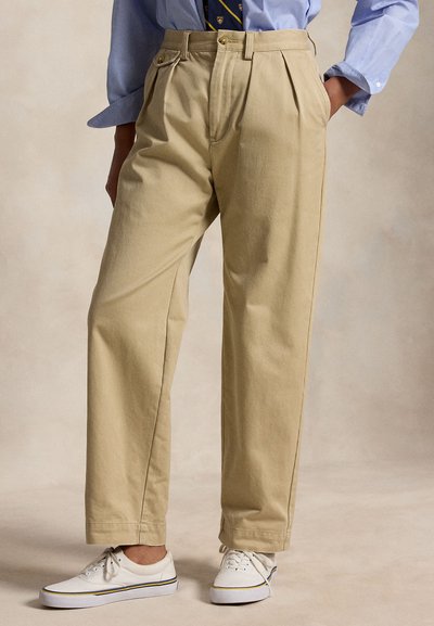 Persoon die beige broek met wijde pijpen, blauwe overhemd, gekleurde das en witte veterschoenen draagt, staat met handen in de zakken.