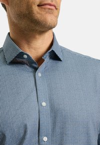 Mann, der ein blau-weißes Karomuster-Hemd mit Kragen trägt, das Kinn und Hals zeigt, mit leichtem Gesichtsstoppel.