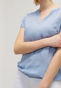 Blusa a maniche corte azzurro chiaro con scollo a V, caratterizzata da una leggera fantasia bianca. Tessuto morbido con una vestibilità comoda.