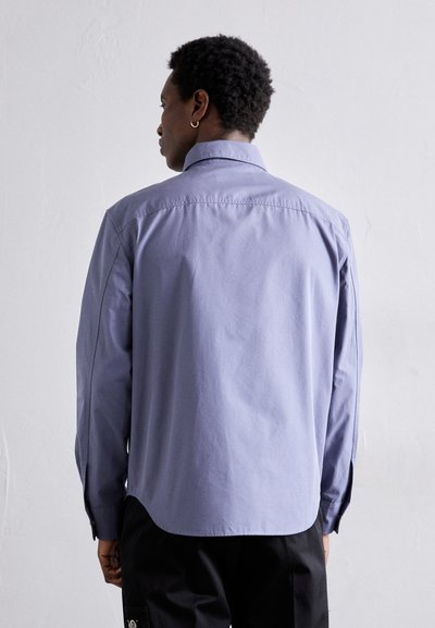Camisa de manga larga color púrpura claro con cuello, textura suave, dobladillo ligeramente curvado y puños abotonados. Presenta un diseño simple y limpio.