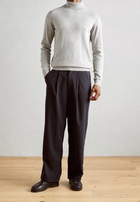 Lysegrå rullekrave-trøje kombineret med mørke bukser med plisseringer og nålestriber samt sorte lædersko, sat mod en neutral baggrund.