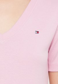  Ροζ βαμβακερό πουκάμισο με V-λαιμόκοψη και κοντά μανίκια. Διαθέτει ένα μικρό ναυτικό μπλε, κόκκινο και λευκό λογότυπο στην περιοχή του στήθους. Λείος υφασμάτινος ιστός.