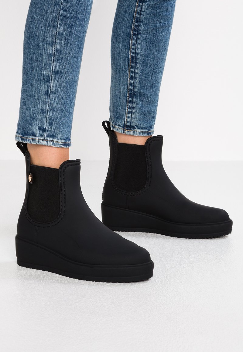 Bottines noires en caoutchouc, présentant un empeigne courbée et des panneaux latéraux élastiques. La semelle est plate avec une texture antidérapante.