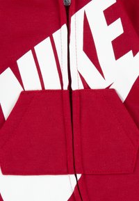 Κόκκινη φούτερ ζακέτα με φερμουάρ από βαμβάκι, με μεγάλο λευκό λογότυπο "NIKE", δύο μπροστινές τσέπες και αντίθετους λευκούς δίσκους που ενσωματώνουν γεωμετρικό σχέδιο.