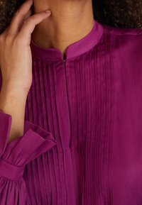 Lila pusero, jossa on pleat-tyylisiä yksityiskohtia, korkea kaulus ja löysät hihat, joissa on kerättyjä hihansuita. Sileä, kevytyhytli.