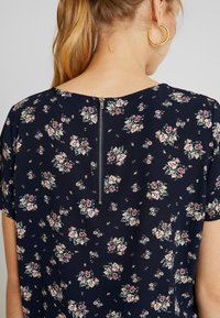 Marinebluse mit floralem Muster, die über einen runden Ausschnitt, kurze Ärmel und einen sichtbaren Reißverschluss im Rücken verfügt. Der Stoff hat eine glatte Textur mit rosa und grünen Blumenmustern.