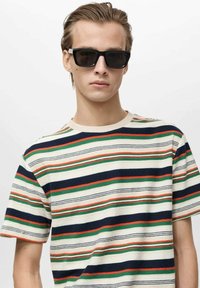 T-shirt rayé crème, avec des rayures horizontales en marine, vert et rouge. Col rond, manches courtes, texture douce. Lunettes de soleil noires portées.