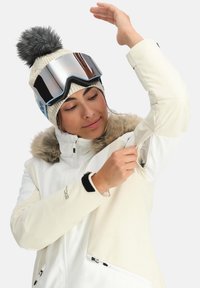 Giacca da sci bianca con colletto in pelo sintetico, polsini regolabili e aperture per le ascelle con zip. Indossata con un cappello di maglia e occhiali a specchio.