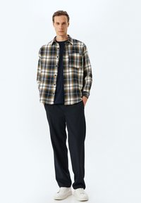 Geknoopte flanellen overhemd met een geruit patroon in crème, marineblauw en groen; gecombineerd met zwarte broek en witte sneakers.