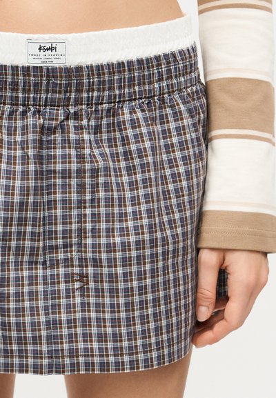 Κοντινό πλάνο μιας καρό φούστας με ελαστική μέση σε καφέ, μπλε και λευκό χρώμα, με λευκή εσωτερική ταινία και ένα χέρι που ακουμπά δίπλα της.