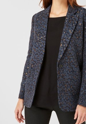 Donna che indossa un blazer blu e nero a fantasia con un bottone singolo sopra una maglietta nera semplice e pantaloni neri, in piedi con le mani rilassate.