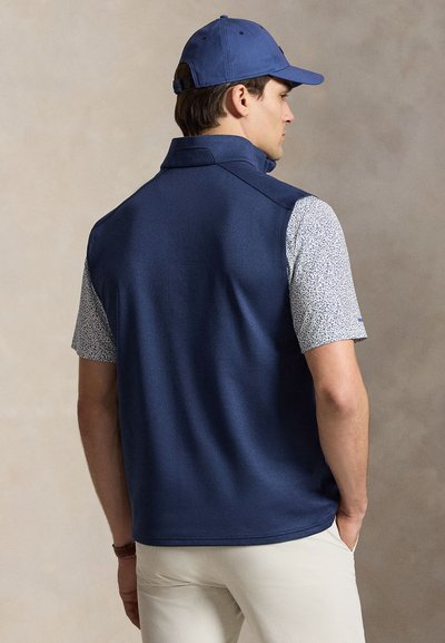 Homme portant une casquette bleu marine, un gilet bleu marine, une chemise blanche à manches courtes avec motif et un pantalon de couleur claire, vu de dos, les mains dans les poches.