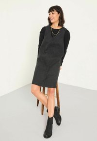 Dunkelgraues Knopfverschluss-Kleid aus strukturiertem Stoff mit figurbetontem Design, kombiniert mit einem schwarzen Langarmoberteil und Ankle Boots.