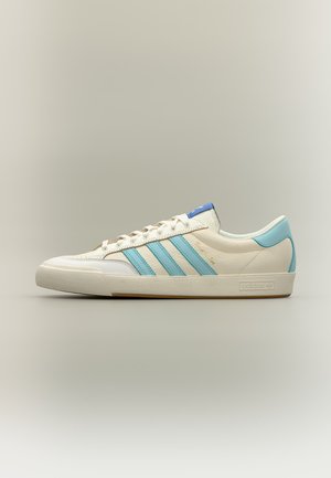 Zapatilla baja de color crema con rayas azul claro de Adidas y lengüeta en el talón, cierre con cordones en la parte delantera y suela plana sobre fondo neutro.