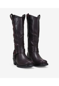 Botas de cuero marrón hasta la rodilla con una textura suave, cremallera lateral y correas en los tobillos. Tacón ligeramente elevado y diseño de punta redondeada.