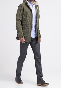Green, hooded jacket with light brown trim and two front pockets. Worn over a light blue shirt and paired with dark jeans and black boots.