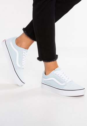 Person trägt hellblaue und weiße Sneakers mit weißen Schnürsenkeln und schwarze Hosen mit ausgefransten Säumen vor weißem Hintergrund.