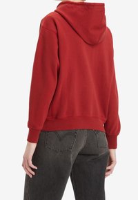 Sudadera roja con capucha, de manga larga, puños acanalados y corte relajado. Llevada con jeans de tiro alto en gris oscuro que presentan un bolsillo trasero con forma de corazón.