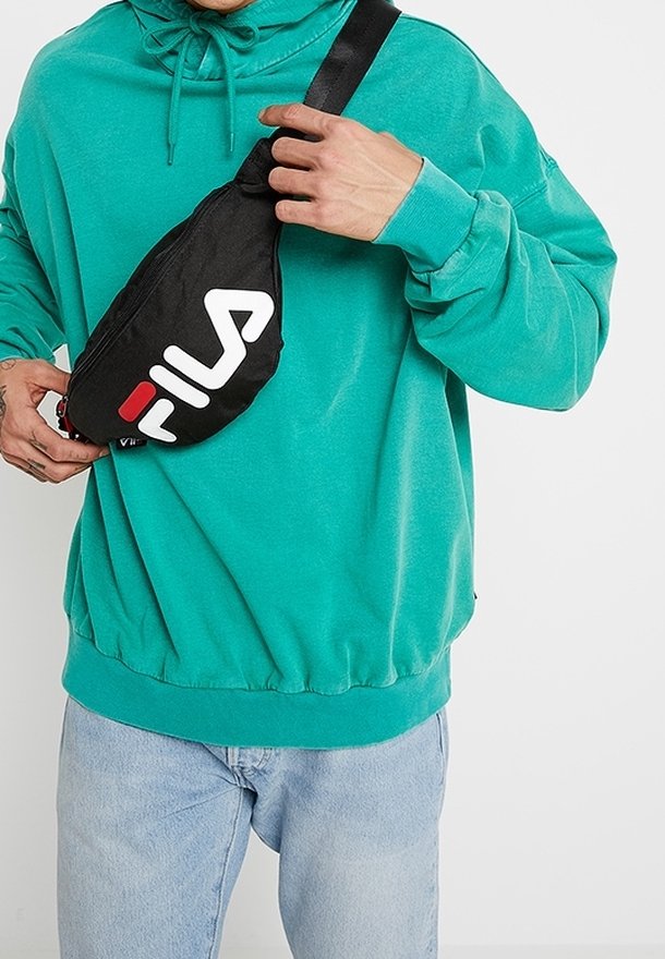 Personne portant un sweat à capuche bleu sarcelle et un jean bleu clair ajustant un sac bandoulière noir Fila sur la poitrine.