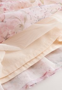 Gelaagde zachte stoffen met bloemmotieven en doorschijnende mesh-texturen in bleke roze en beige tinten, met nadruk op delicate stiksels en materiaaldetails.