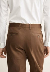 Pantaloni marroni realizzati in tessuto testurizzato, con una vestibilità slim. Due tasche posteriori con bottoni, indossati con una camicia bianca.