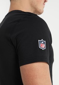 Camiseta de algodón negra con mangas cortas. Parche del logo de la NFL destacado en color en el hombro izquierdo. Textura suave y ajuste clásico.