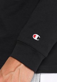 Schwarzes Langarmshirt aus Baumwolle mit gerippten Bündchen; hat ein kleines gesticktes Logo in Weiß, Rot und Blau am Ärmel.