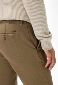 Bruine linnenmix broek met een gladde textuur, voorzien van een knoopzak aan de achterkant en een elastische tailleband. Een lichtgrijze geribbelde mouw van een trui is zichtbaar.