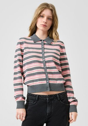 Jonge vrouw draagt een grijs, roze en wit gestreepte cardigan met knopen en lange mouwen en zwarte high-waisted jeans tegen een effen achtergrond.