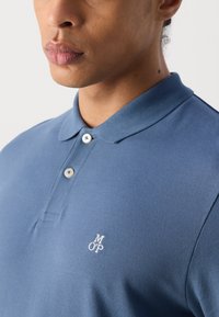 Cămașă polo albastră dintr-un material texturat, cu guler, două butoane și un logo brodat în alb pe pieptul stâng.