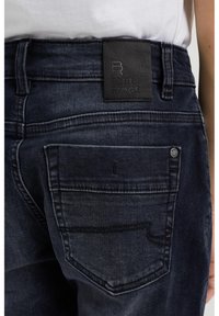 Tmavě modré džíny z denimu s texturovaným povrchem, s černou koženou páskou na opasku s vyraženým nápisem "BLUE RIDGE" a detaily na zadní kapse.