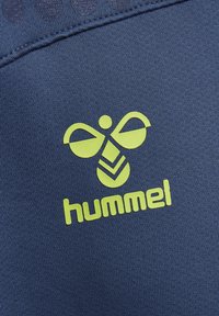Neongeel Hummel-logo met een gestileerde bij boven de merknaam op een navyblauwe, gestructureerde stofachtergrond.