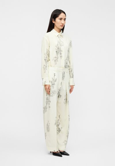 Εκρού σύνολο με μαύρα λουλουδάτα γραμμικά σχέδια, που περιλαμβάνει πουκάμισο με κουμπιά και παντελόνια με φαρδιά πόδια, ολοκληρωμένο με μυτερές μαύρες γόβες.