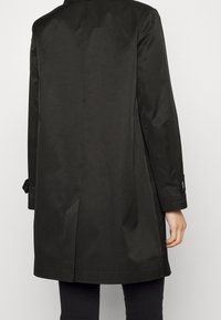 Svart knälång kappa med en mitt bak-springa, knappade manschetter, bärs av en person som vänder ryggen mot kameran med sin högra hand synlig.