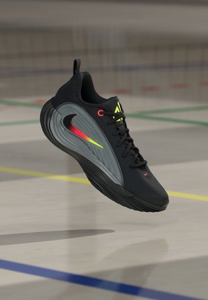 Chaussure de sport noire et grise avec un swoosh Nike coloré, des lacets noirs et des accents jaunes, flottant au-dessus d’un sol de gym avec des lignes de terrain.