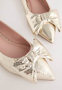 Auksinės spalvos netikro odos butukai su smaigaliu, turintys tekstūruotą žvynelio raštą ir išskirtinę didelę kaspinio detalę priekyje.