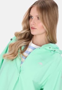 Menta zöld vízálló dzseki állítható kapucnival, cipzárral és logó részlettel. Világoskék-fehér csíkos ingre viselve.
