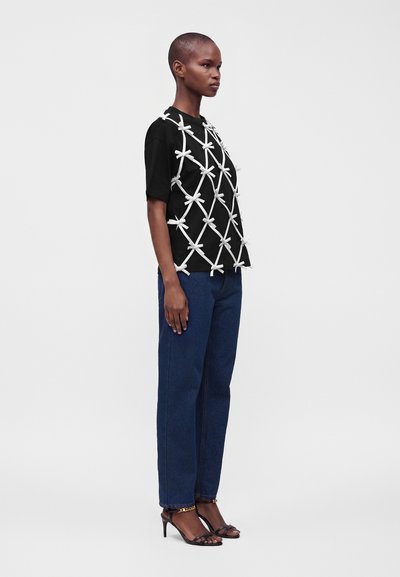 Femme aux cheveux courts portant un t-shirt noir avec de grands nœuds blancs, un jean bleu et des sandales noires à talons hauts, debout de profil.
