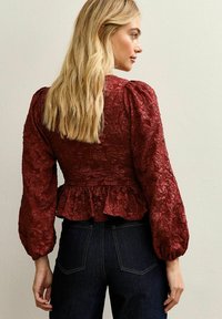 Blusa roja texturizada con mangas abullonadas, dobladillo péplum y un intrincado patrón floral. Combinada con jeans de tiro alto en azul oscuro.
