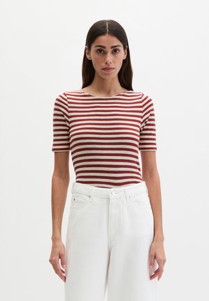 Femme portant un haut à manches courtes rayé rouge et blanc et un pantalon blanc taille haute, debout devant un fond blanc uni.