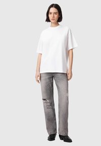 Young woman standing wearing a plain white oversized T-shirt, gray ripped straight-leg jeans, and black ankle boots against a neutral background.