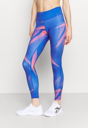 Leggings bleus avec un motif géométrique rose et violet, dotés d'une taille haute et d'une texture lisse et extensible, associés à des baskets blanches.